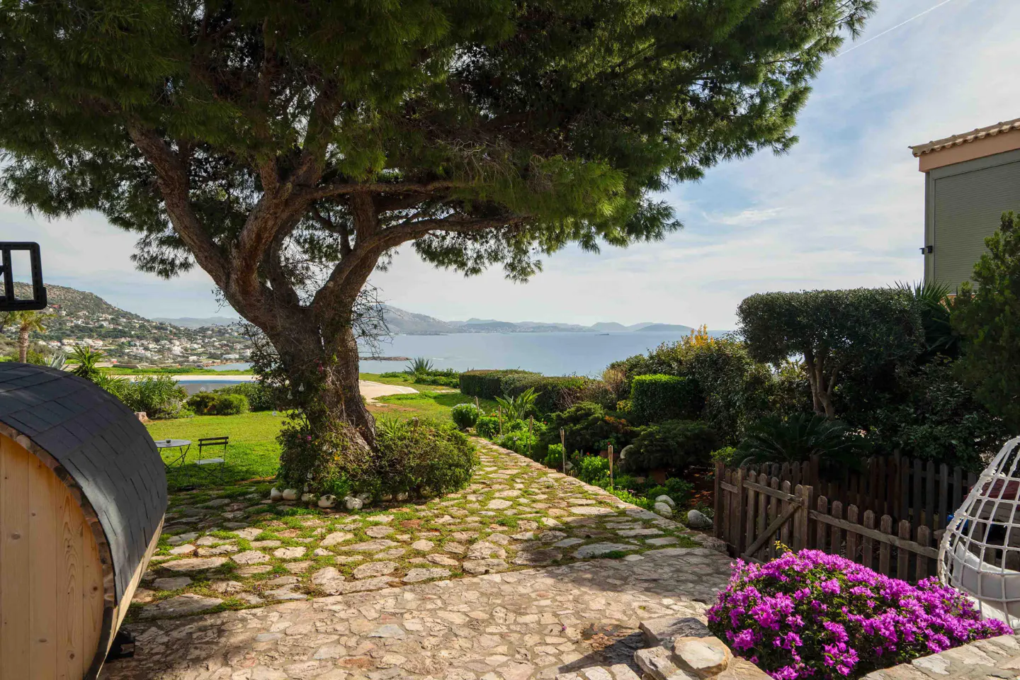 Landscaped backyard with stone path leading to ocean view. A large tree provides shade. Purple flowers and a wooden sauna are in the foreground.