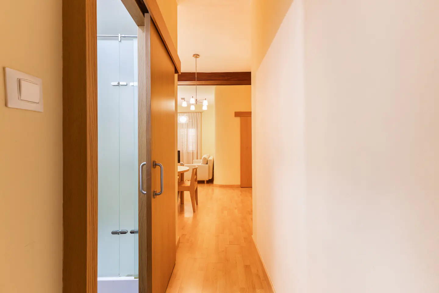 A hallway with light wood floors leads to a bright living area with a dining table and sofa, viewed past a sliding door.