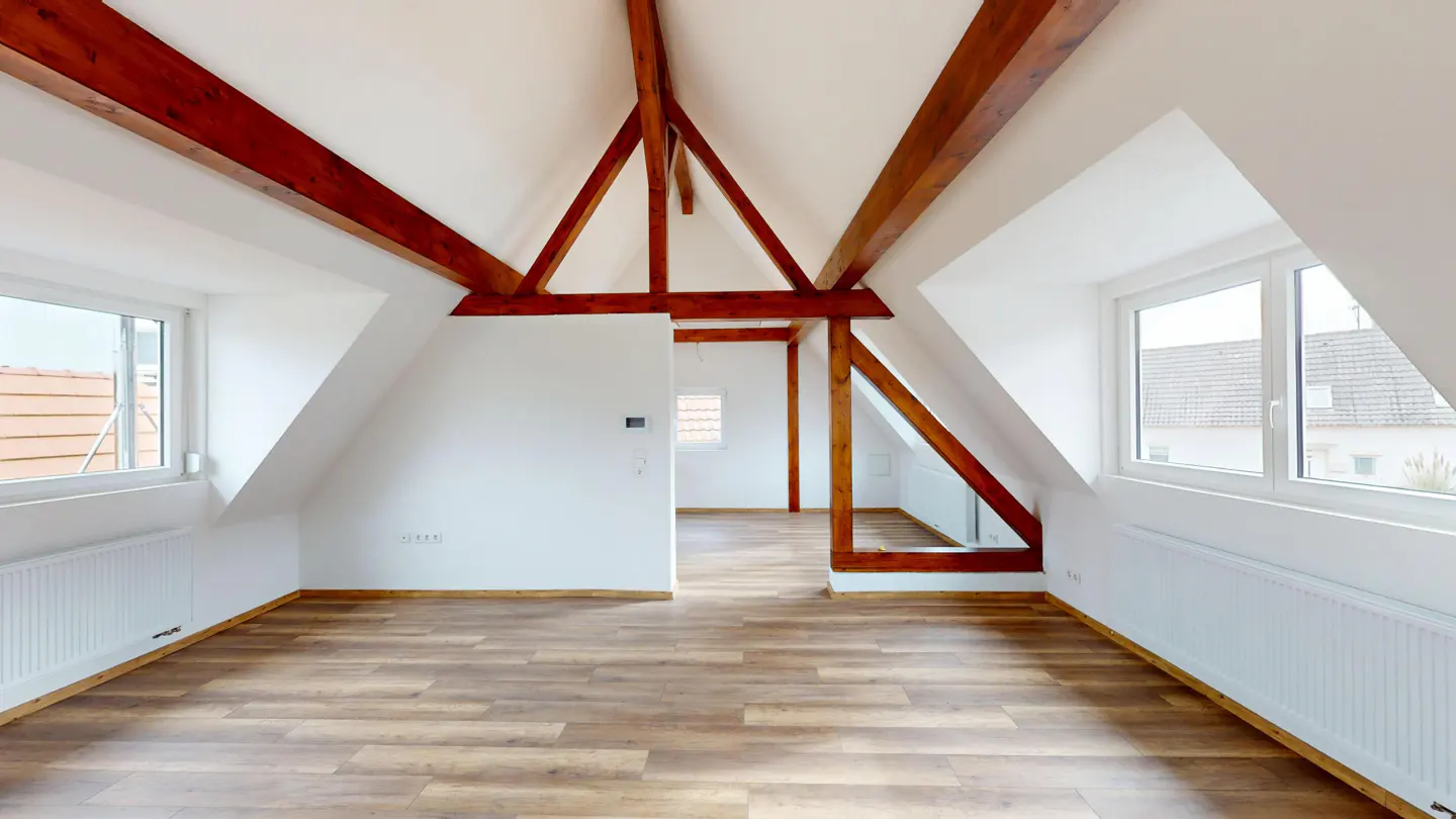 Attic room with wood floors, white walls, and exposed dark wood beams. Two windows let in natural light.