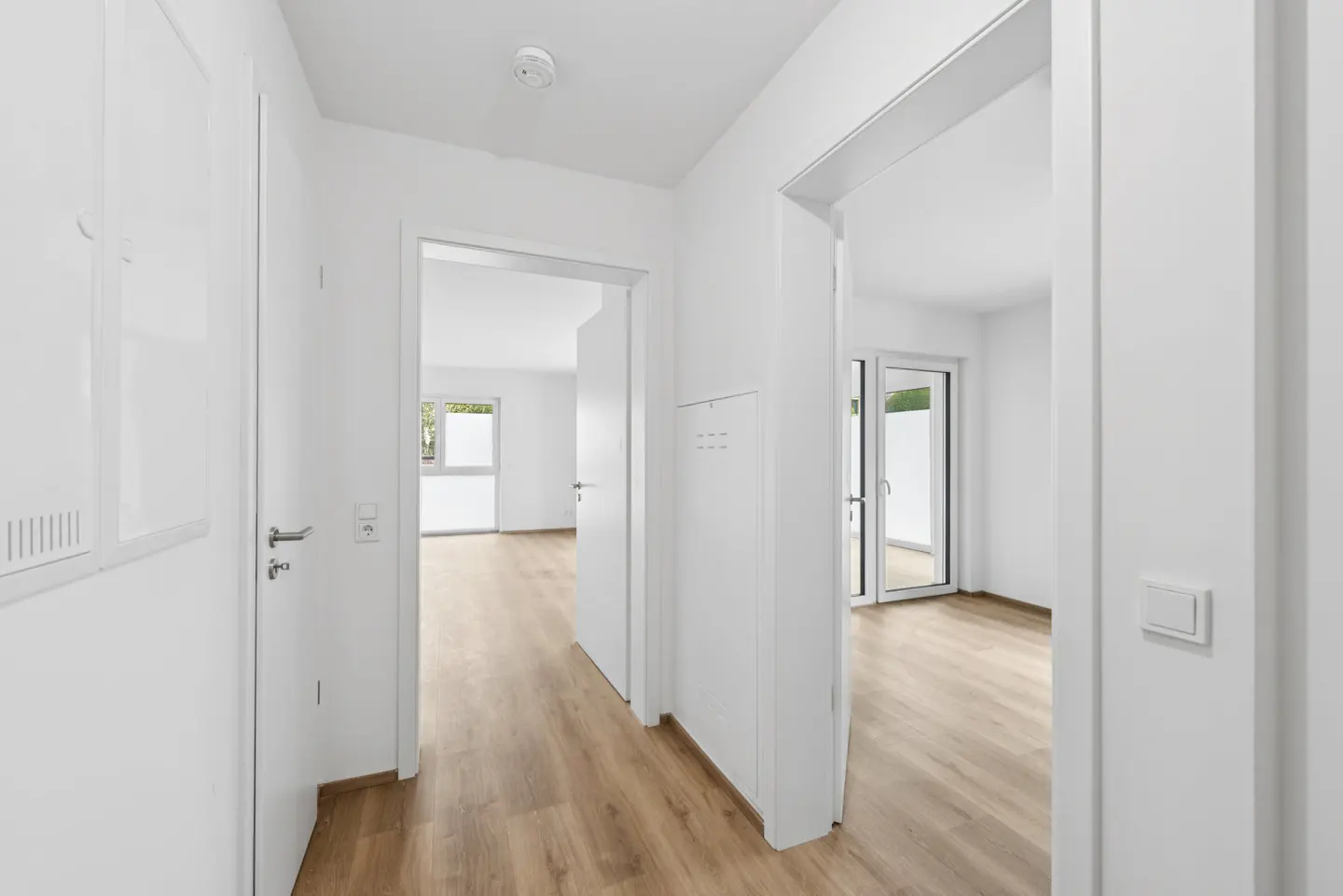 Bright, white hallway with light wood floors. Open doorways lead to rooms with windows and glass doors.
