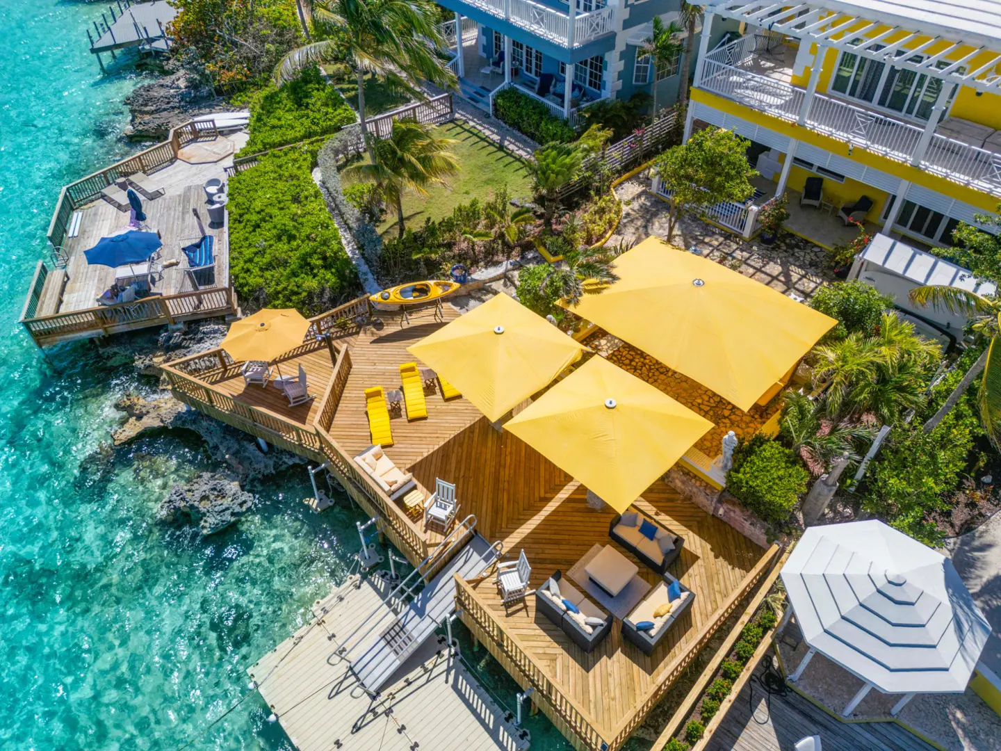 Aerial view of a waterfront property with wooden decks, yellow umbrellas, lounge chairs, and turquoise water. Houses and greenery are in the background.