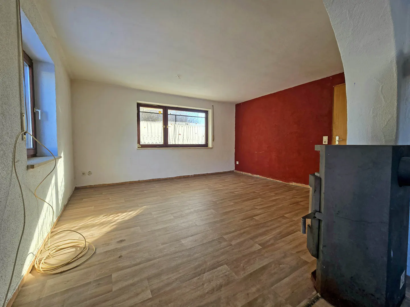 An empty room with wood-look floors, white walls, a red accent wall, and two windows. A black wood-burning stove is in the foreground.
