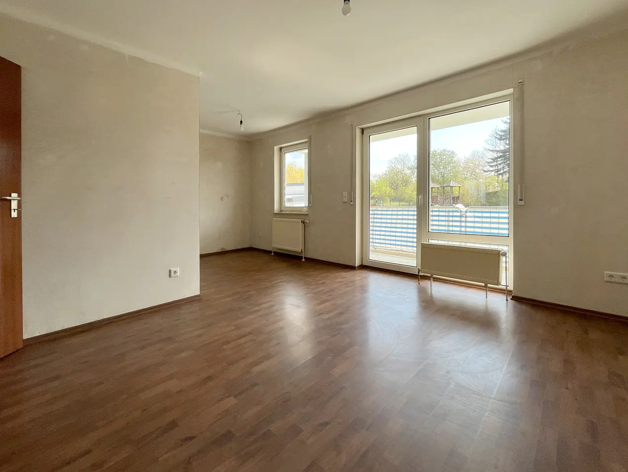 Empty room with brown wood floors, beige walls, and a door on the left. A window and sliding glass doors lead to a balcony with a blue and white railing.