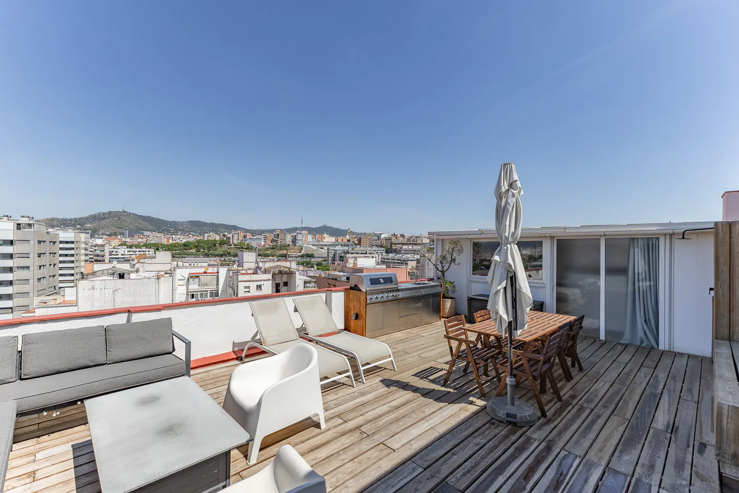 Rooftop terrace with wooden decking, outdoor dining set, lounge chairs, sofa, and a grill, overlooking a city skyline.