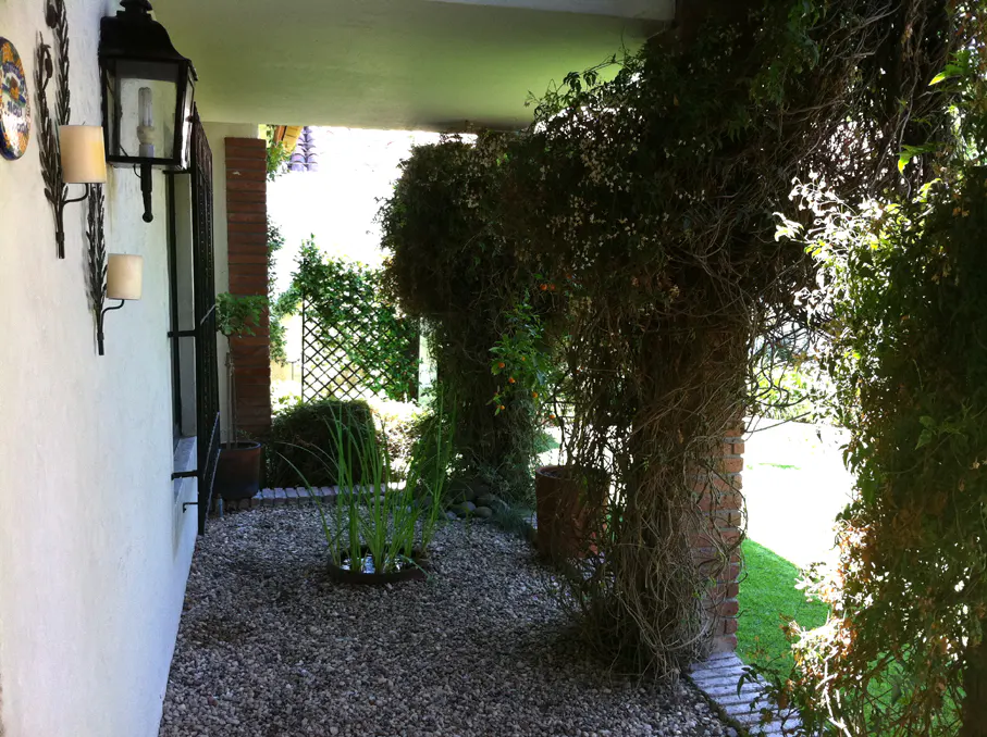 Covered walkway with stone ground, lush greenery, and brick pillars. Wall sconces and a lantern adorn the white wall.