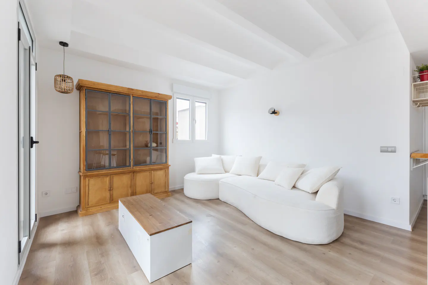 Bright living room with white walls, wood floors, a curved white sofa, a wooden cabinet, and a modern coffee table.