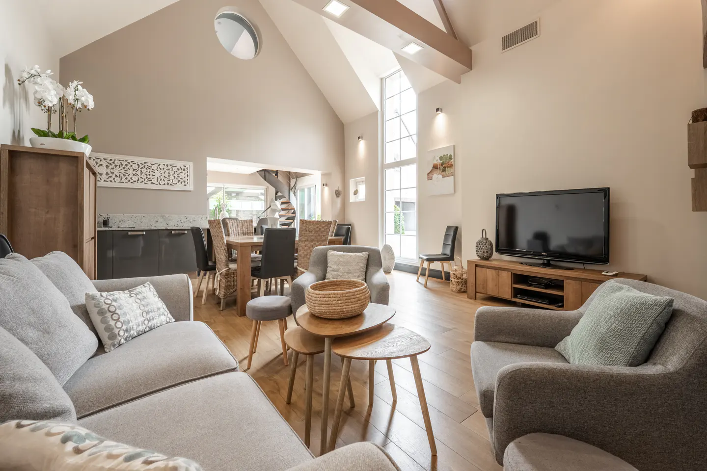 Bright, open living room with gray sofas, wooden tables, and a large TV. A dining area and spiral staircase are visible in the background.