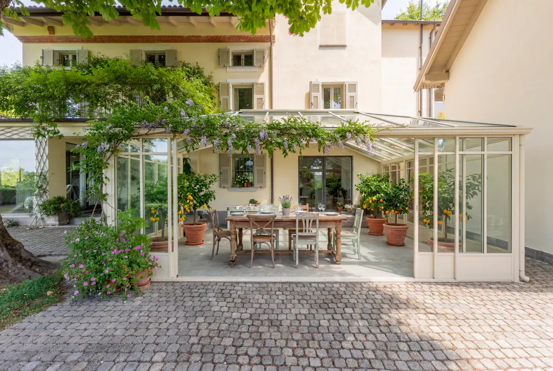 Exterior view of a cream-colored house with a glass-enclosed patio, a wooden table set for a meal, and potted citrus trees.