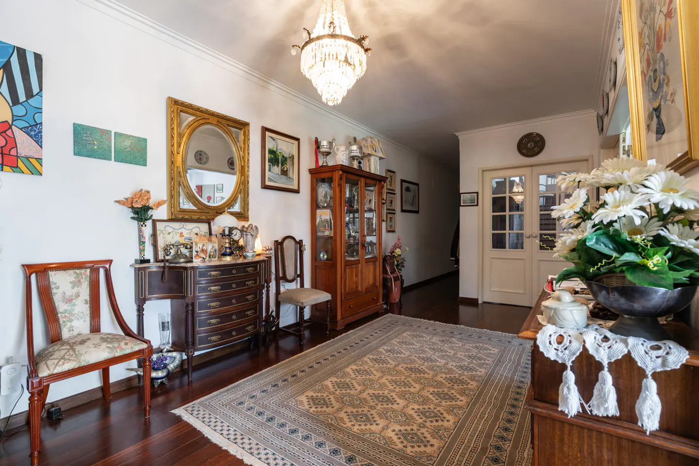 A well-lit foyer with dark wood floors, a patterned rug, and antique furniture under a crystal chandelier.
