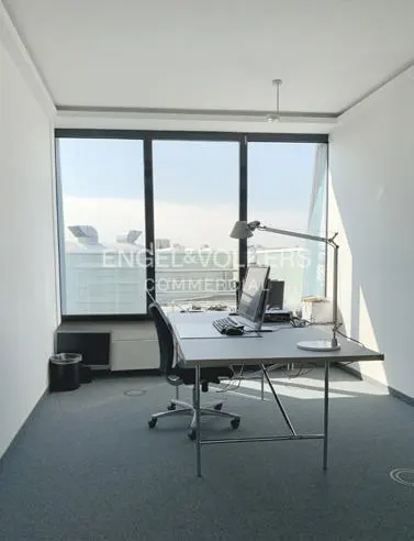 Bright office space with a desk, chair, laptop, and large window overlooking a cityscape. The walls are white, and the floor is gray carpet.