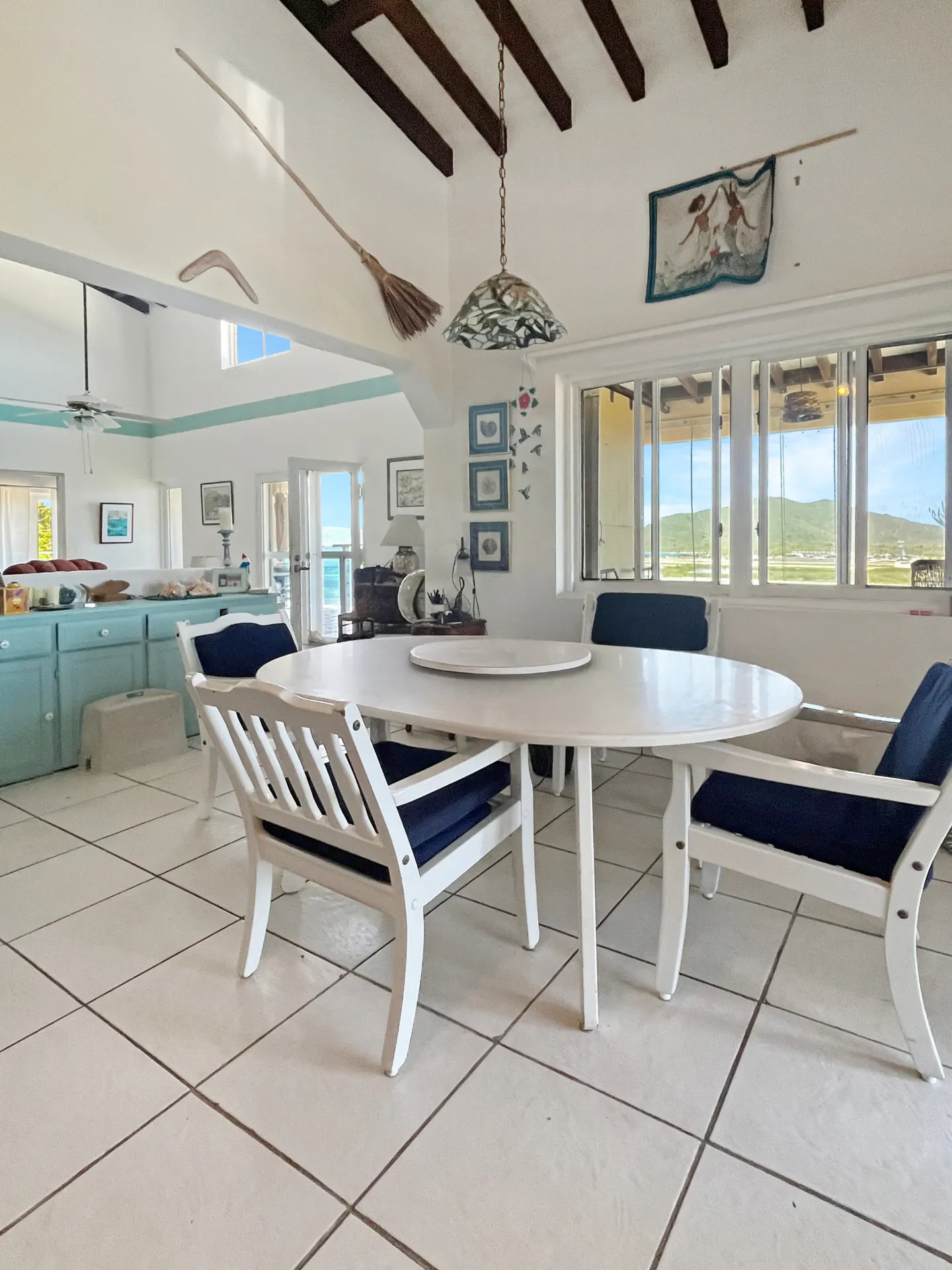 Bright dining area with white table and chairs with blue cushions. Broom decor on ceiling. Ocean view through windows.
