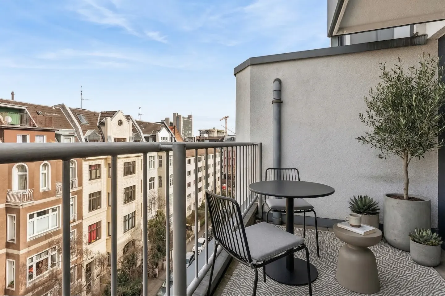 Balcony view with black table and chairs, potted plants, and a patterned rug. City buildings are visible in the background.