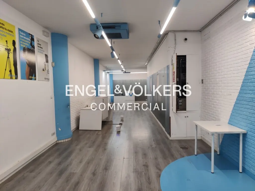 Empty commercial space with gray wood floors, white walls, and blue accents. Fluorescent lights illuminate the room. Engel & Völkers logo is visible.