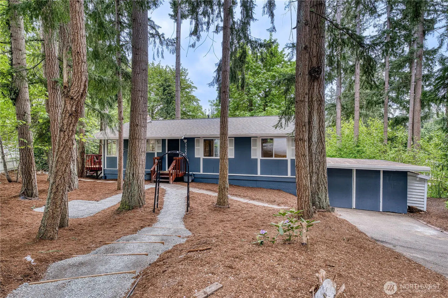 Blue house with white trim, surrounded by tall trees. A gravel path leads to the front door. The house has an attached garage.