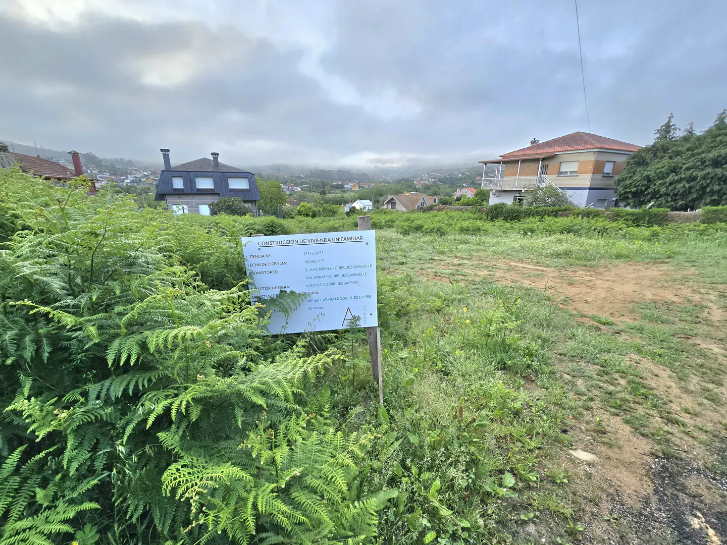 A vacant lot with a construction sign, green ferns, and houses in the background under a cloudy sky.