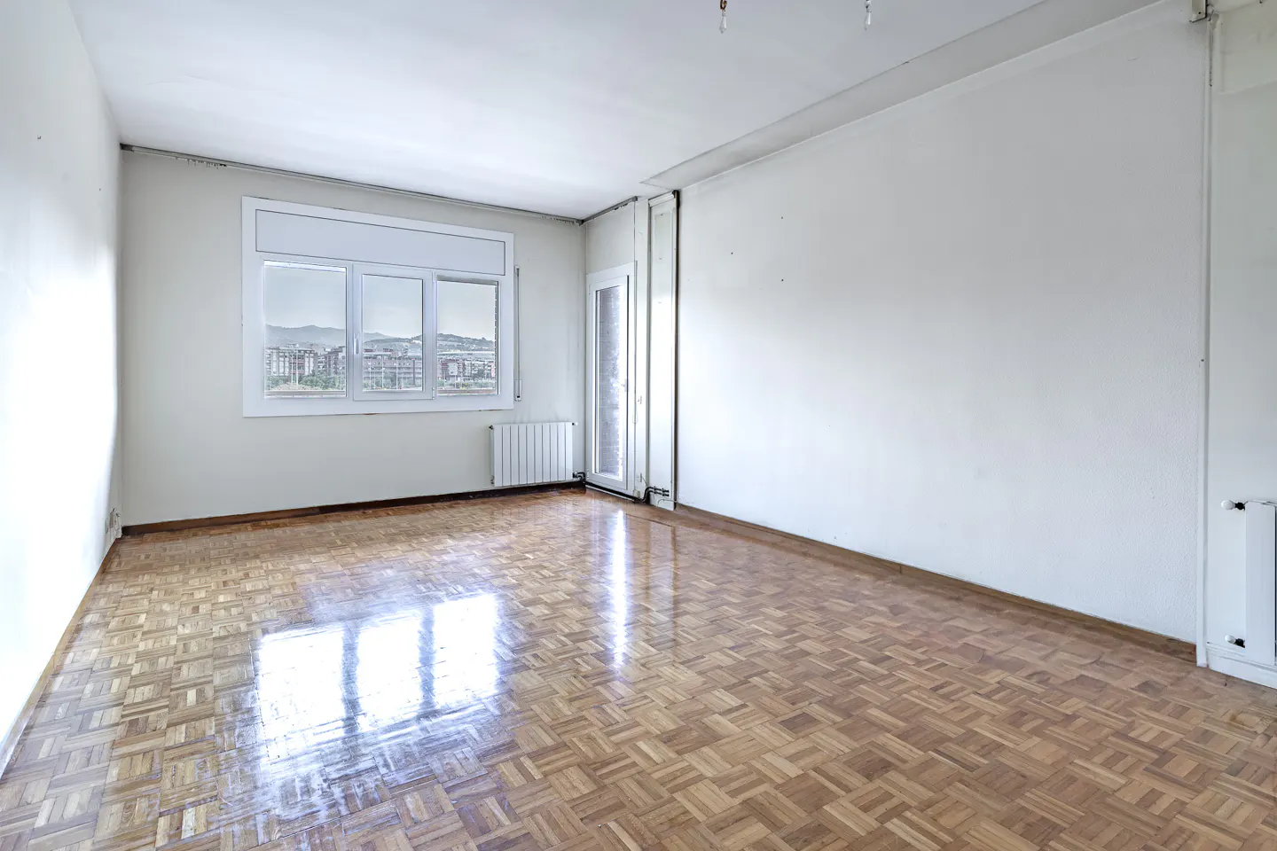 Empty room with parquet floor, white walls, and a window showing a city view.