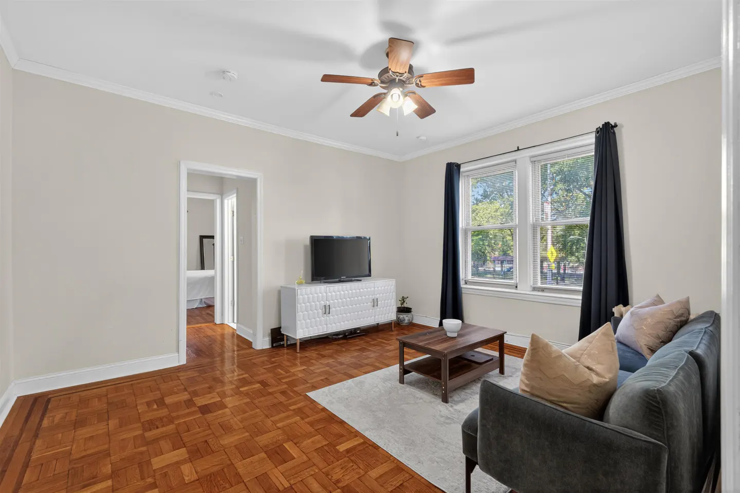 Living room with hardwood floors, a grey sofa, coffee table, white media console with a TV, and windows with black curtains.