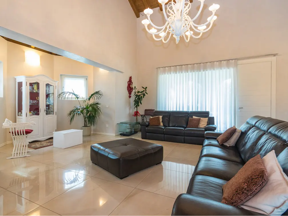 Bright living room with high ceilings, tile floors, and black leather sofas. A white chandelier hangs above. A white cabinet sits in the corner.
