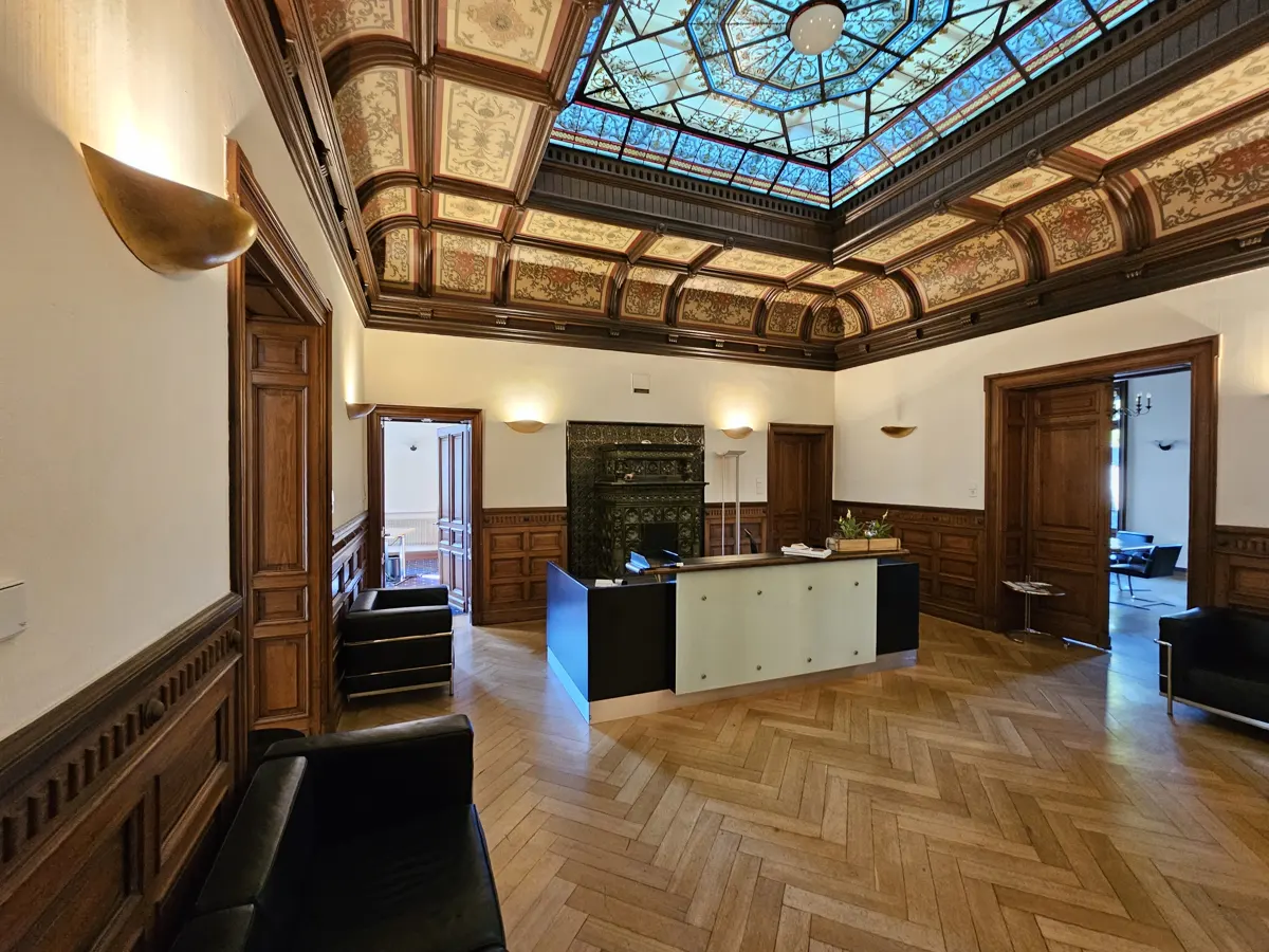 Interior view of a grand lobby with wood paneling, herringbone floors, and a stained-glass ceiling. A reception desk sits in the center.
