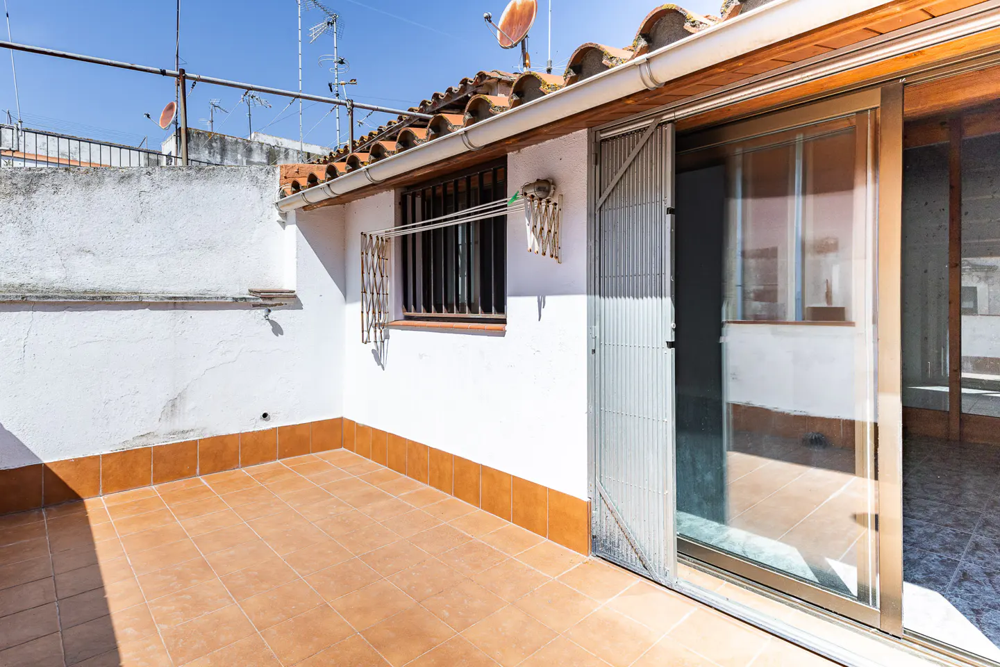 A sunny patio with brown tile flooring and a white wall. A sliding glass door is partially open, and a barred window is visible.