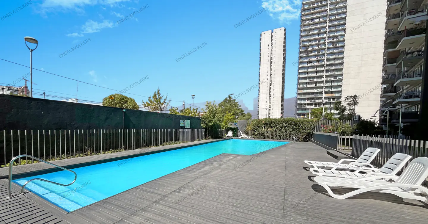 Outdoor pool with lounge chairs on a wooden deck, set against tall buildings and a clear blue sky.