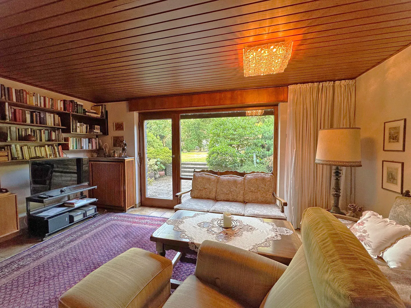 Cozy living room with wood-paneled ceiling, bookshelves, and a large window overlooking a green garden. Furnished with sofas, a coffee table, and a purple rug.