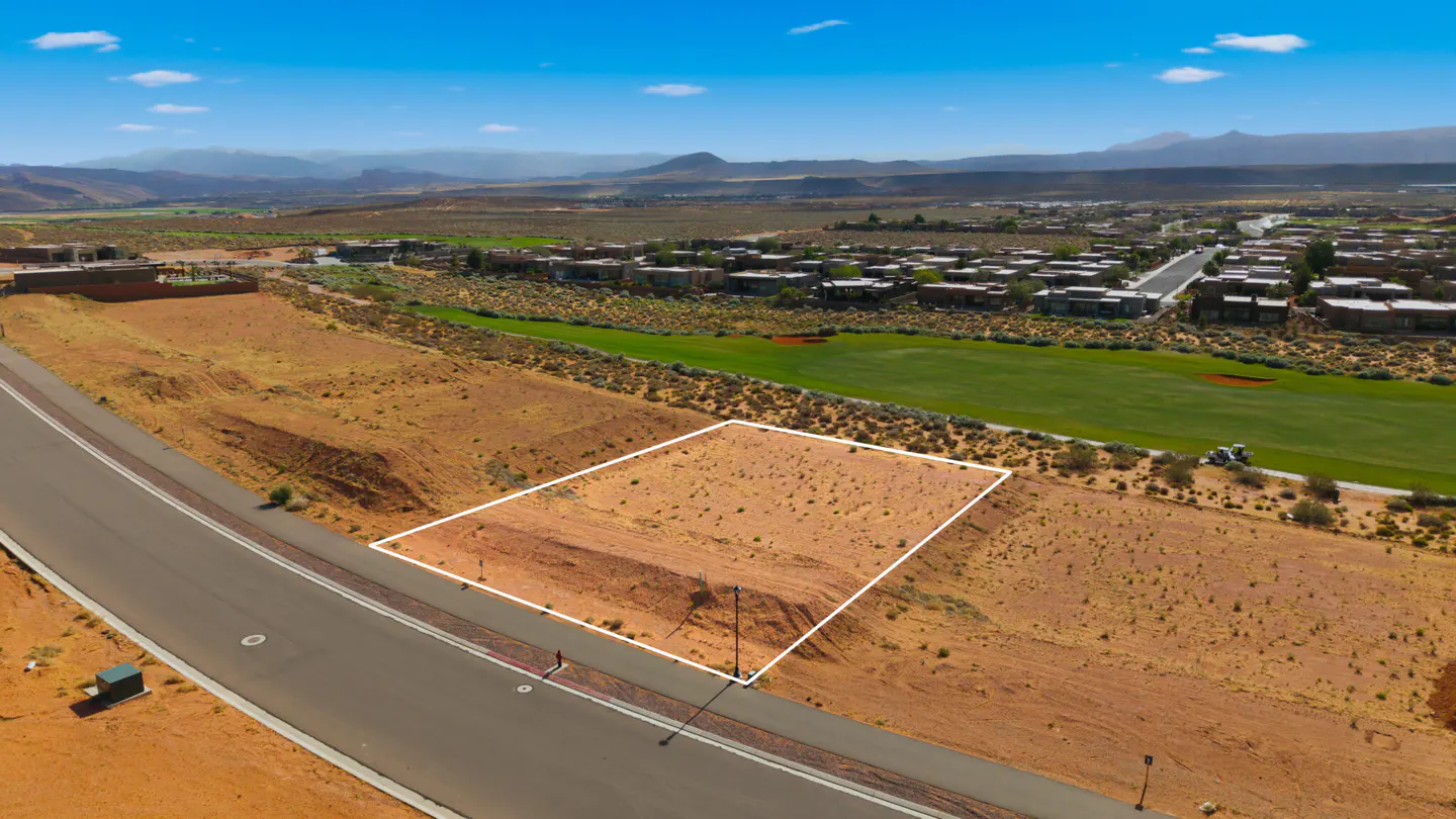 Aerial view of a vacant, rectangular lot outlined in white, adjacent to a road, with a golf course and mountains in the background.
