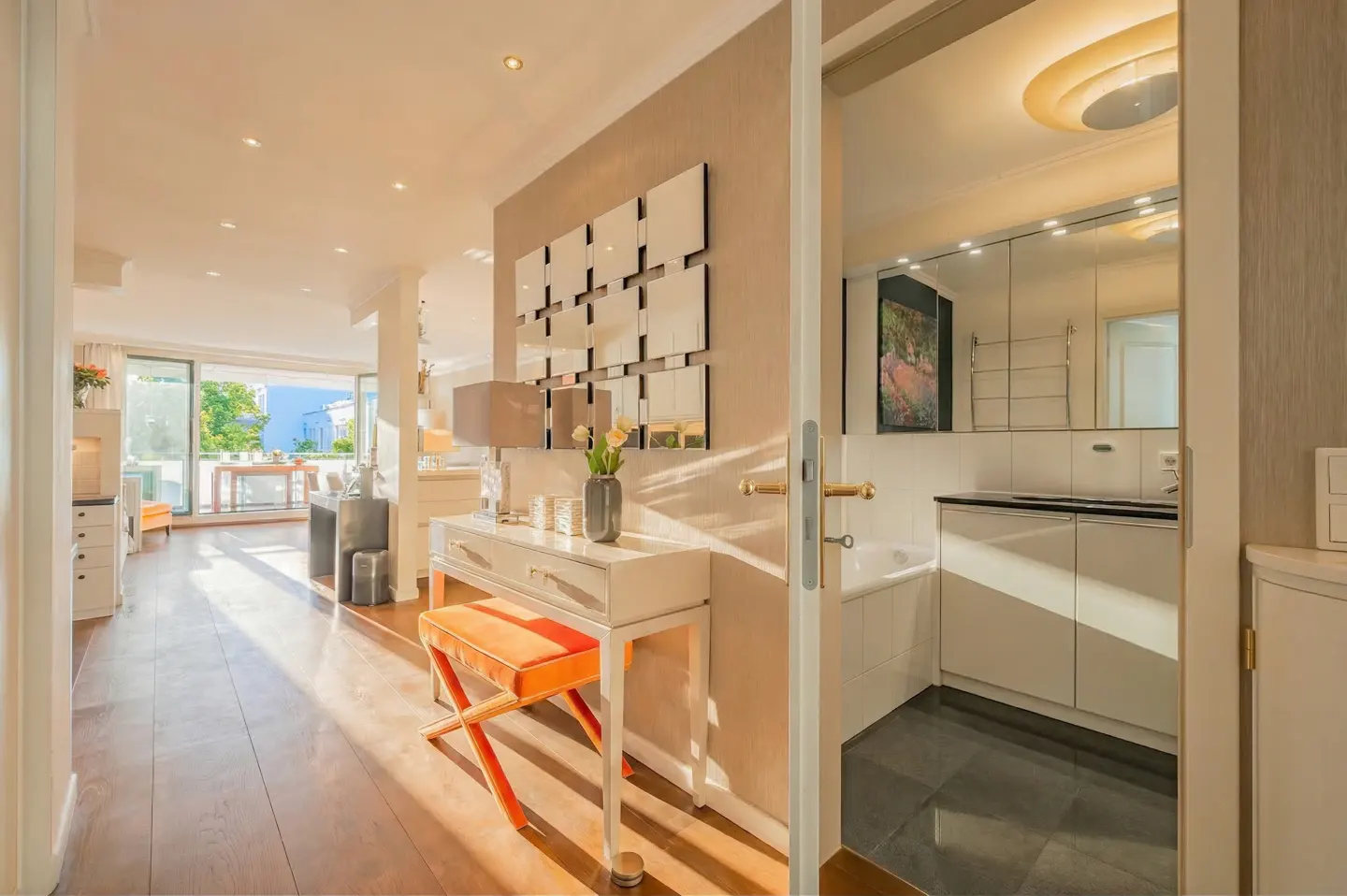 A bright hallway with a white console table, orange stool, and grid mirror, leading to a modern bathroom and living area.