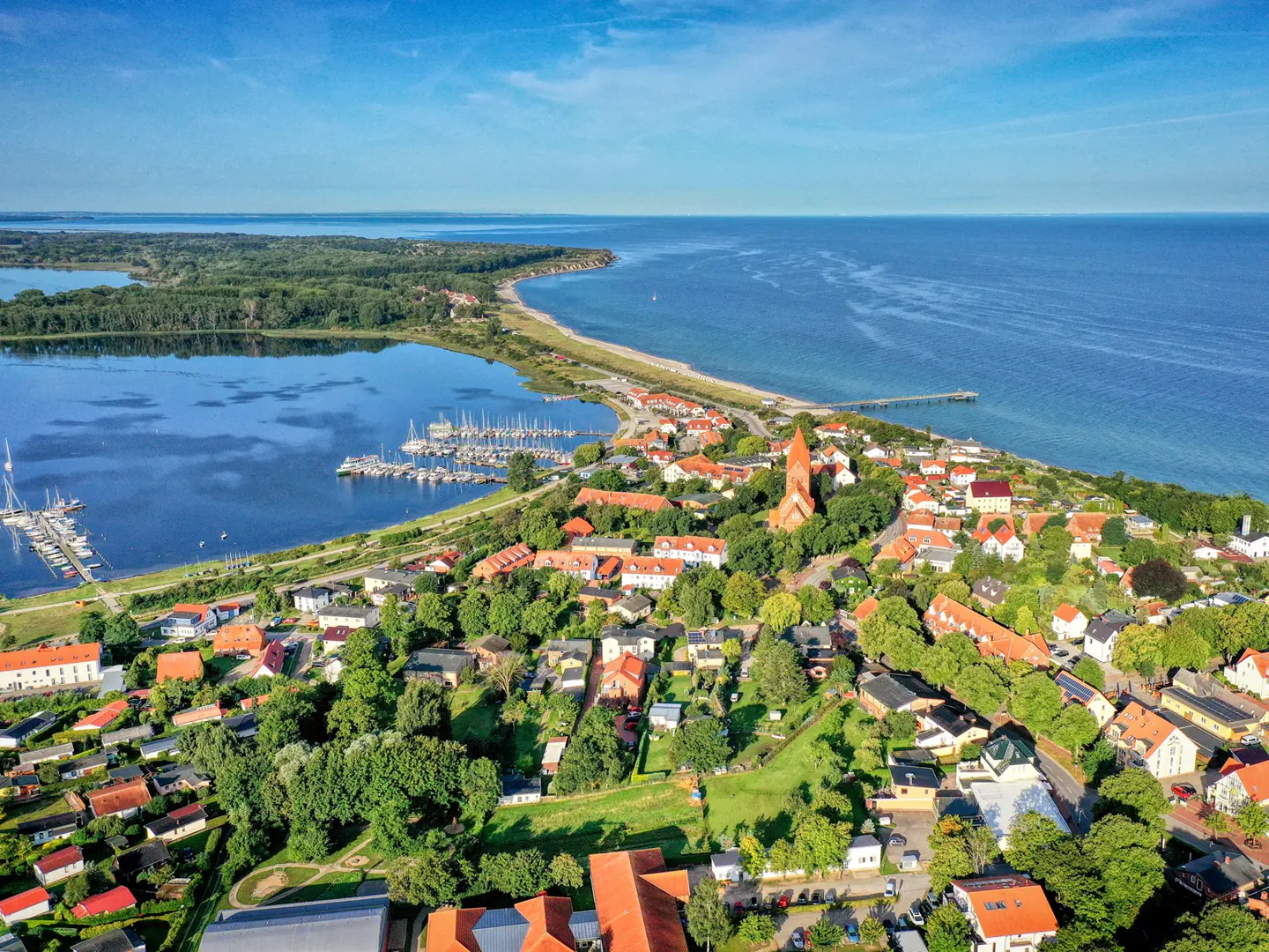 Aerial view of a coastal town with red-roofed houses, a church steeple, and a marina on a blue bay under a clear sky.