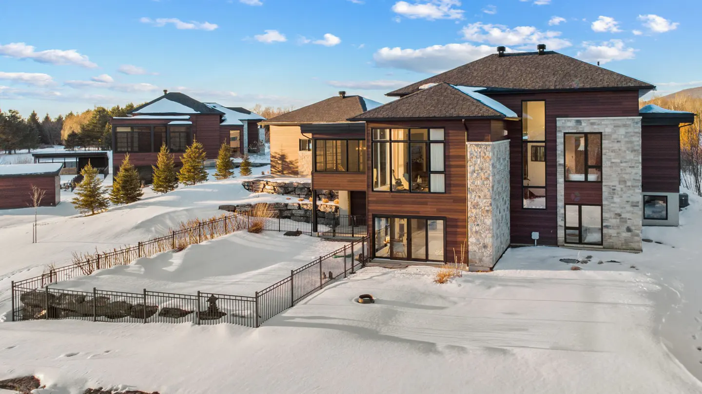 Exterior view of a modern brown and gray house with large windows surrounded by snow and trees under a blue sky.