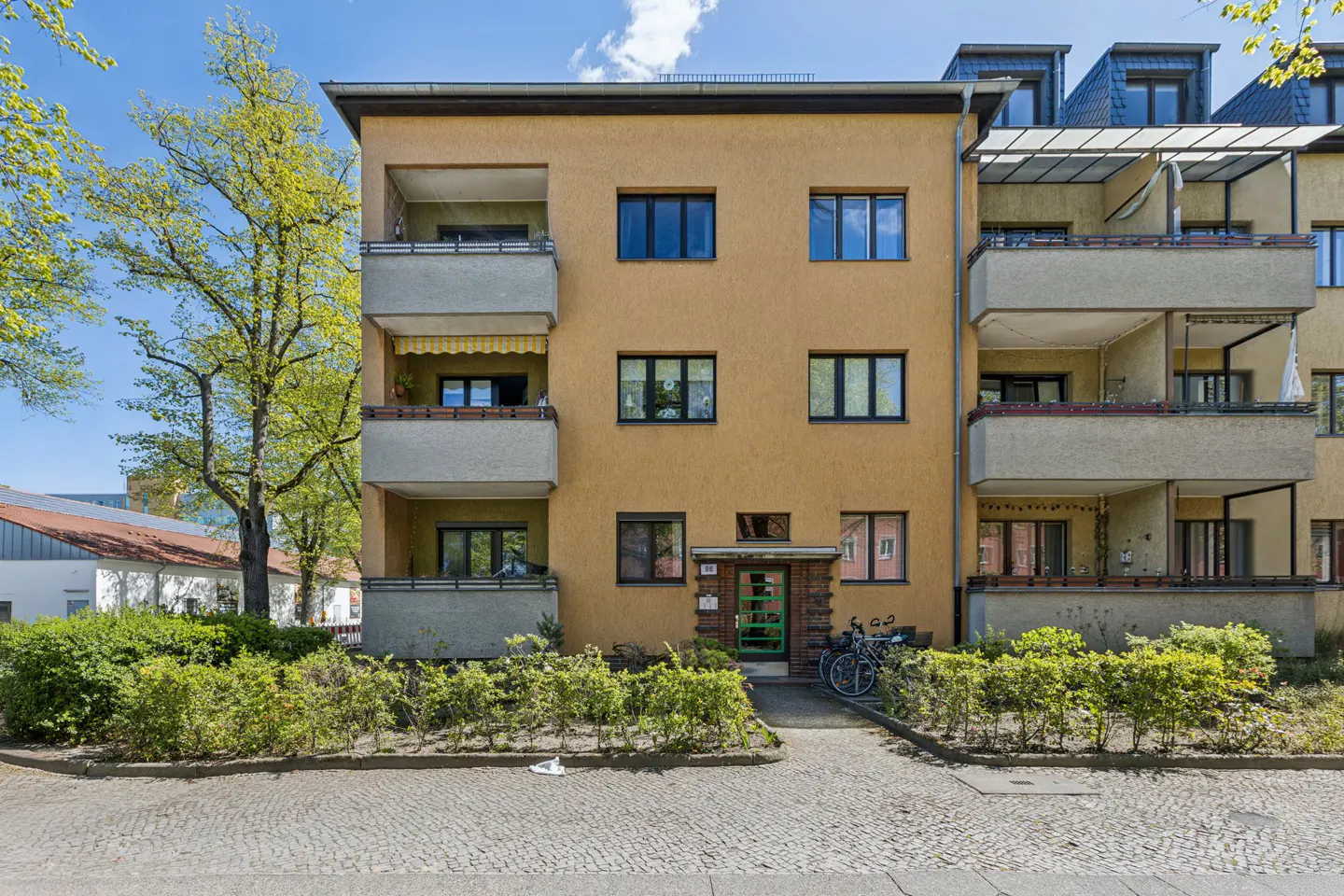A three-story apartment building with balconies, a yellow facade, and dark-framed windows. Bikes are parked near the entrance.