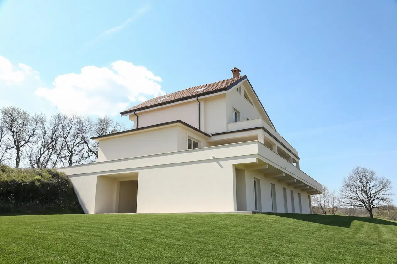 Modern two-story house with a red tile roof and a beige exterior on a green lawn under a blue sky.