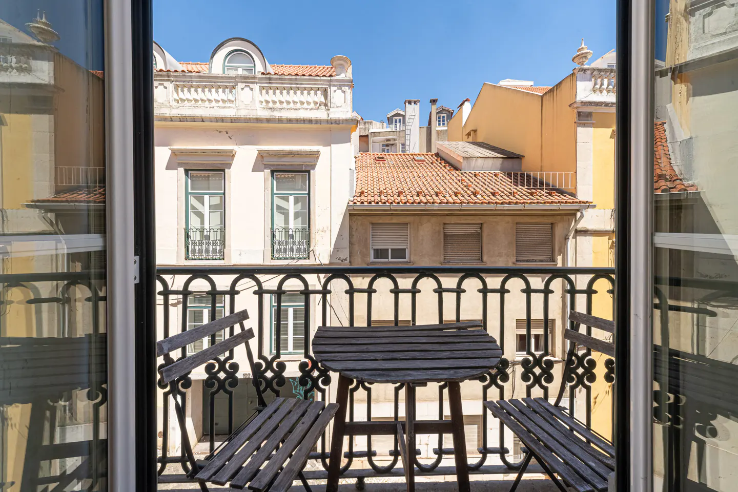 A small balcony with a wooden table and two chairs overlooking historic European buildings with terracotta roofs.