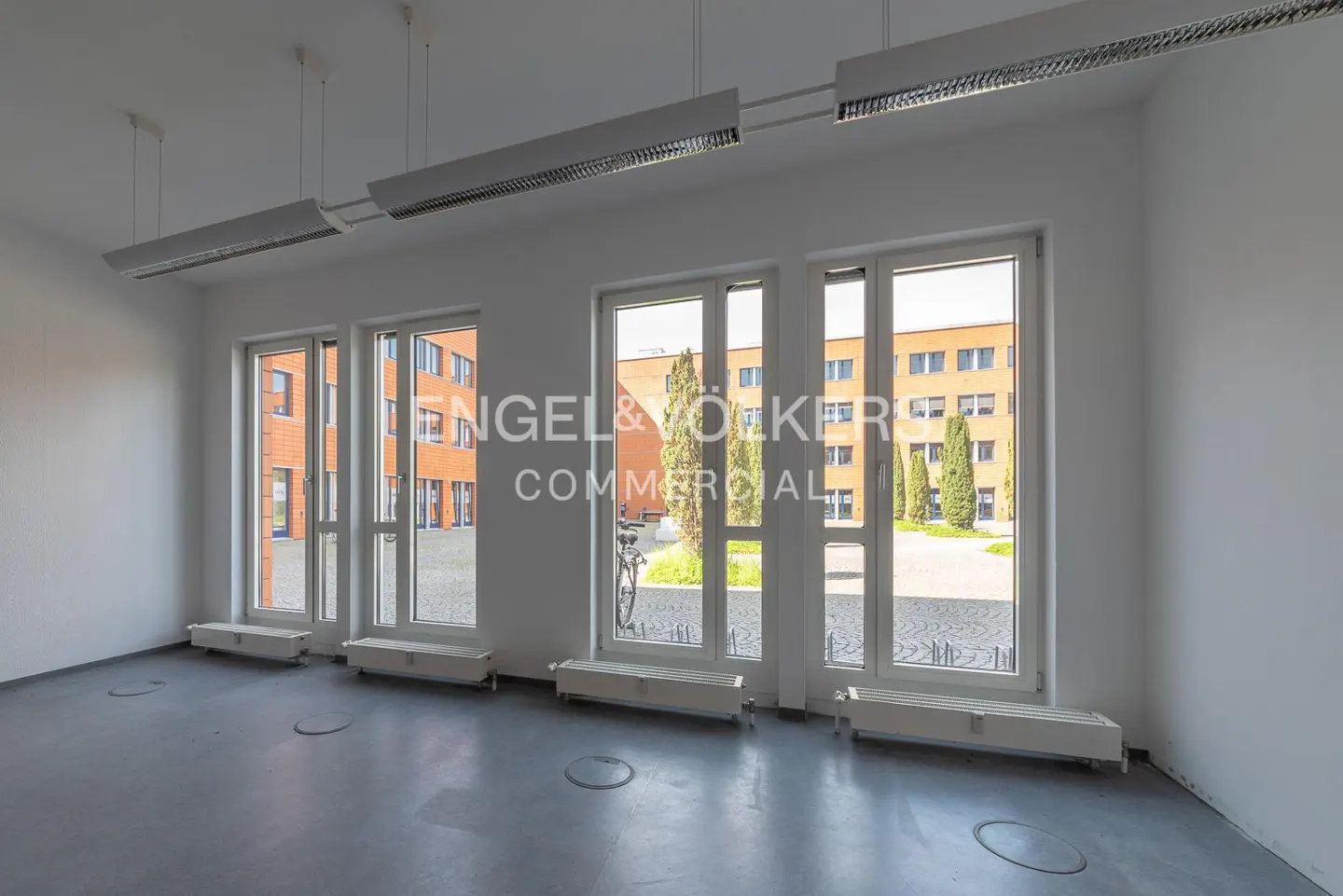 Bright, empty office space with white walls, grey floors, modern ceiling lights, and large windows overlooking a courtyard.