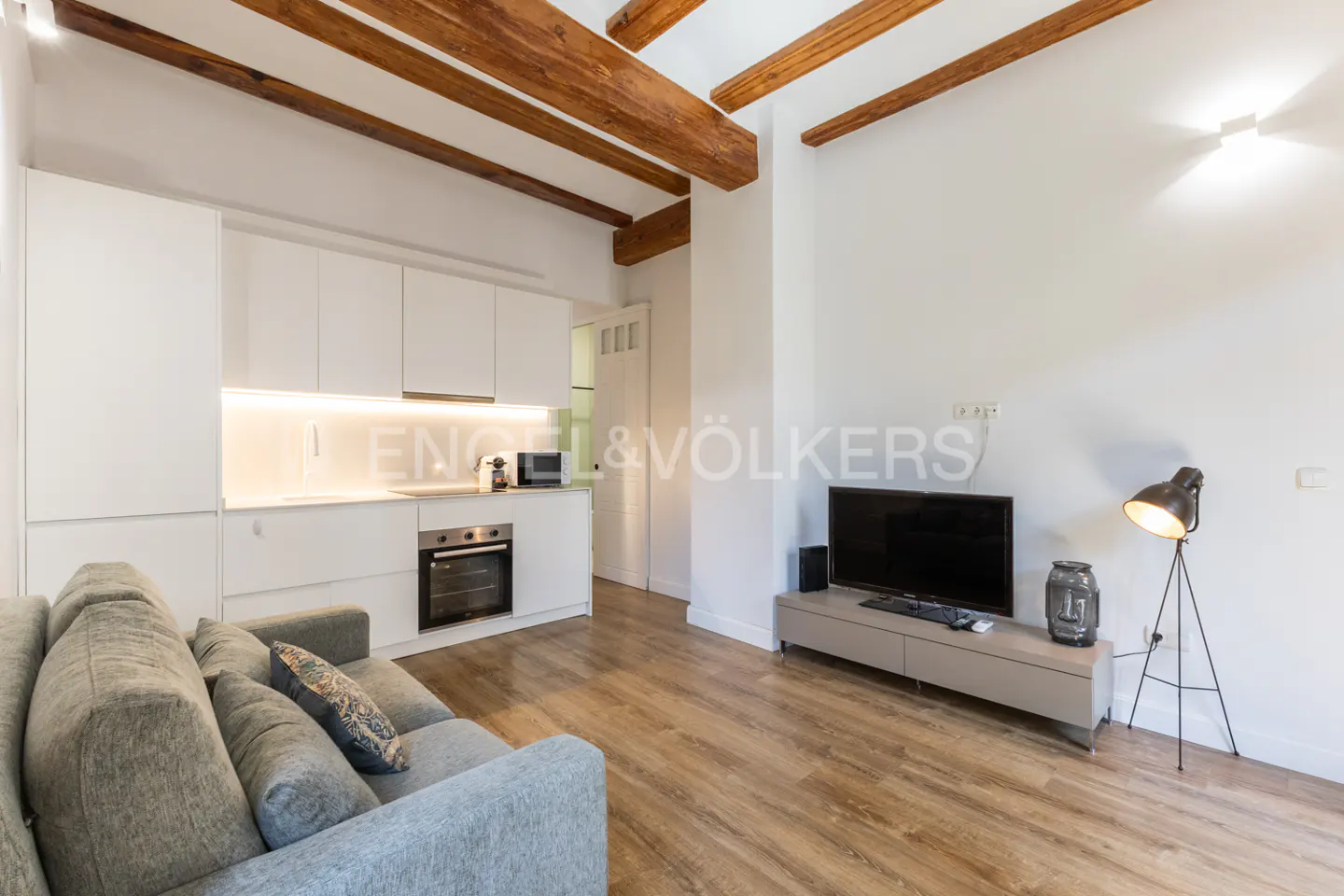 Living room with a gray sofa, white kitchen cabinets, and a TV on a gray stand. Wooden beams are visible on the ceiling.