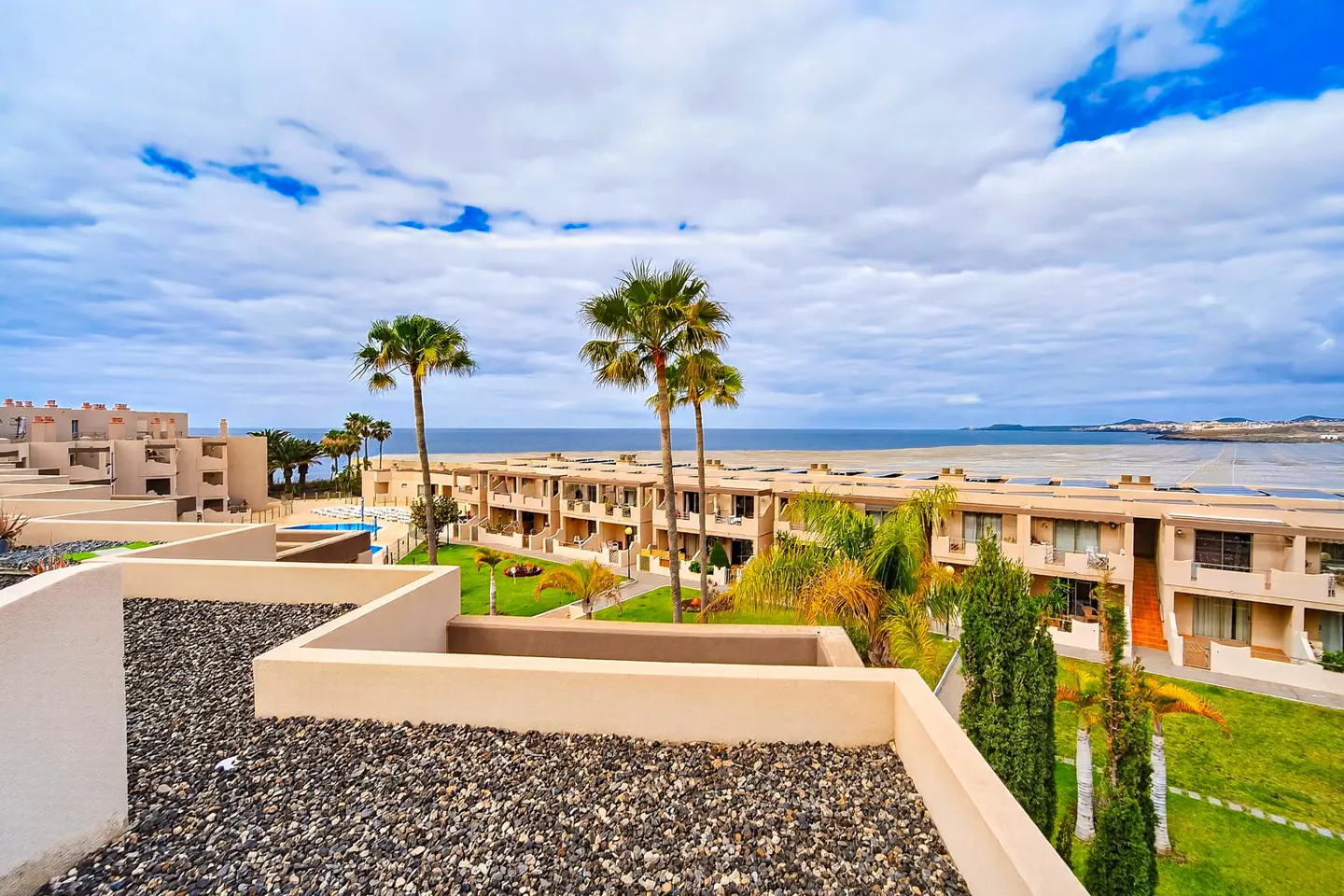 View of a resort with palm trees, beige buildings, and the ocean under a cloudy sky.