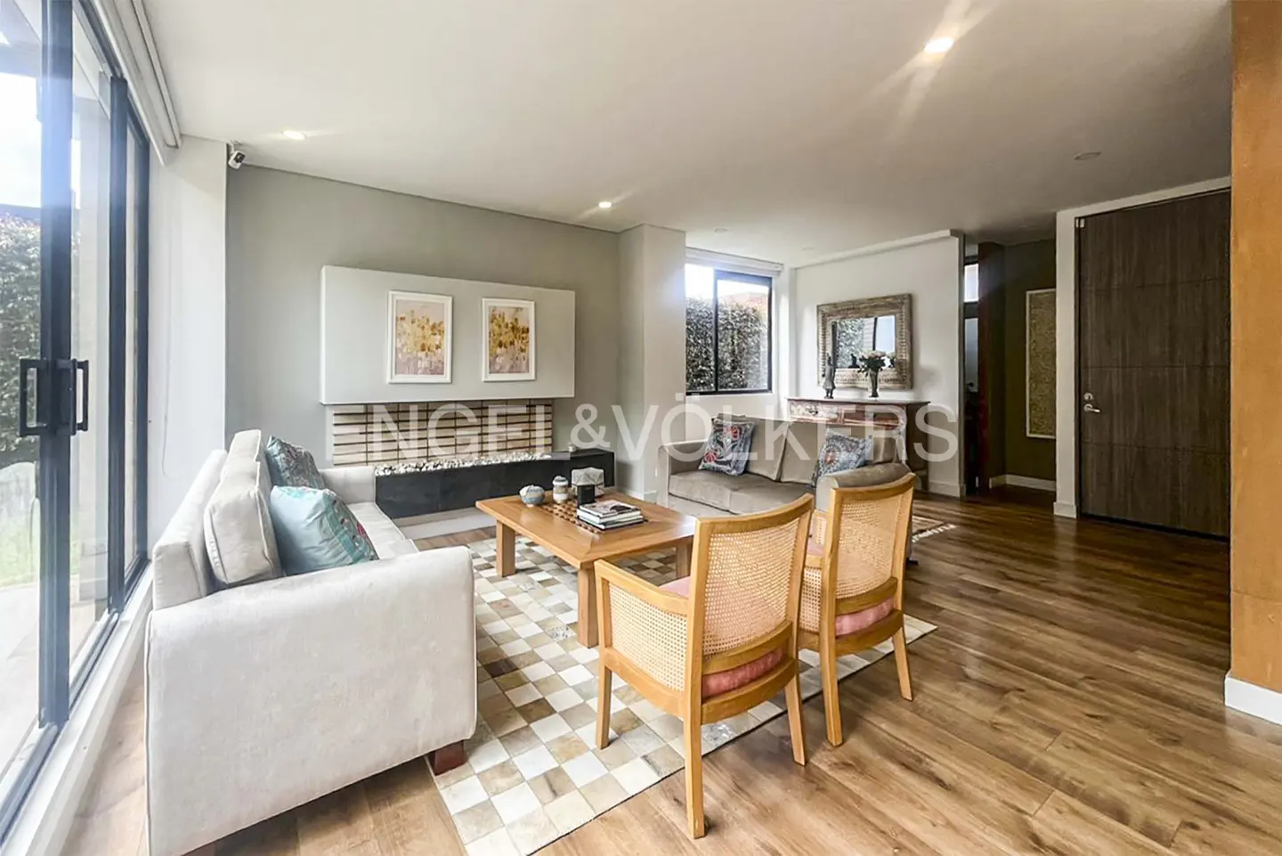 Living room with hardwood floors, a beige sofa, a brown sofa, a wooden coffee table, and two wooden chairs. There is a fireplace with the Engel & Volkers logo above it.