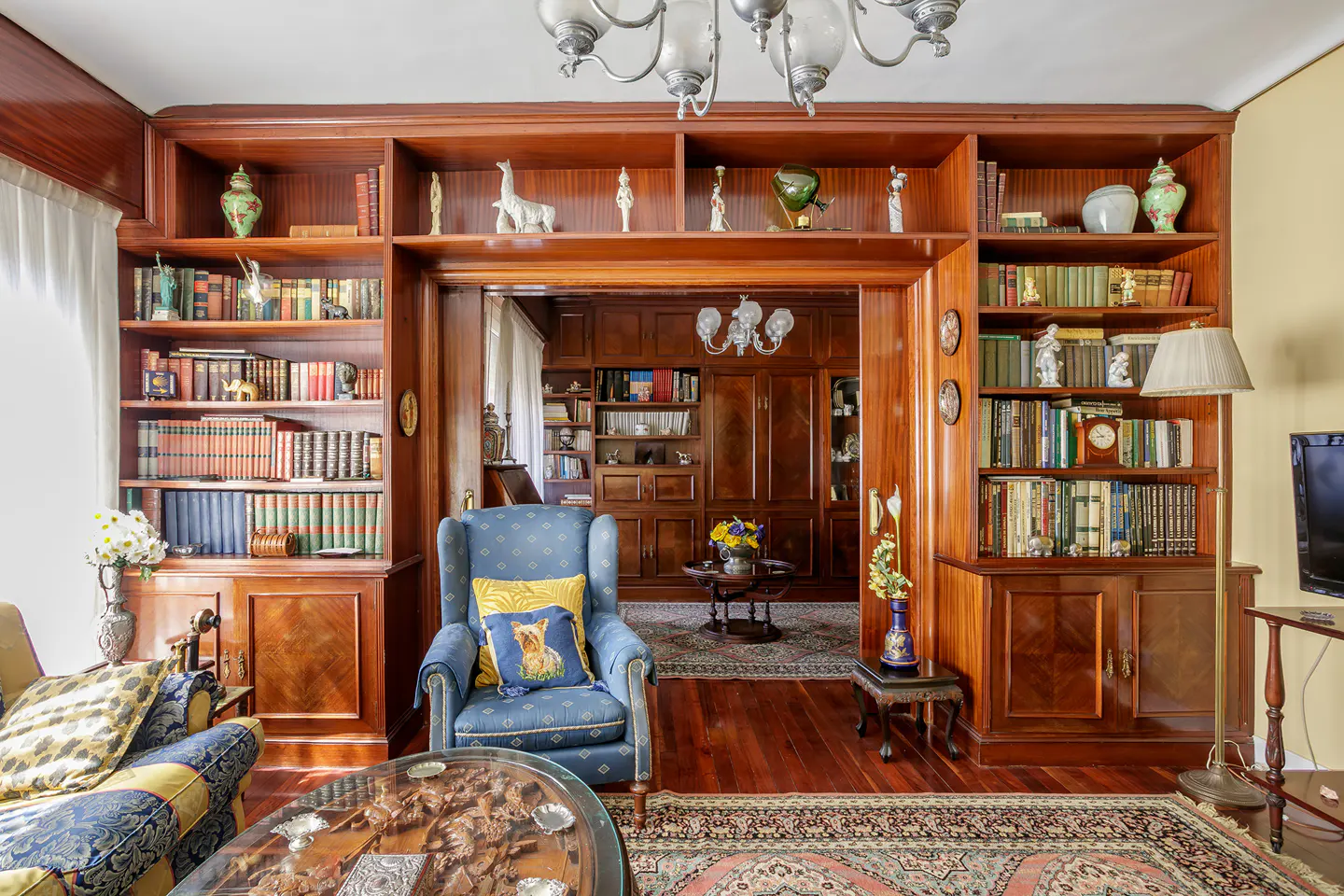 A living room with wood bookshelves filled with books and decorative objects. A blue armchair with a yellow pillow sits near a carved wood coffee table.