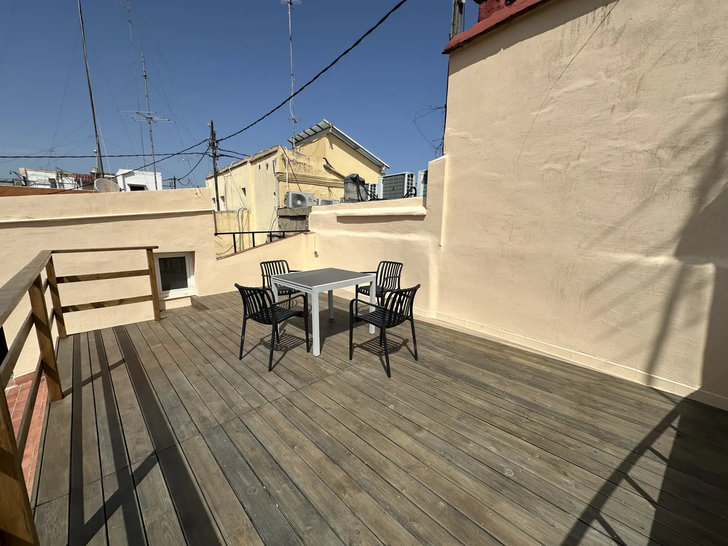 Outdoor patio with wooden deck, table, and four black chairs. Beige walls and blue sky in the background.