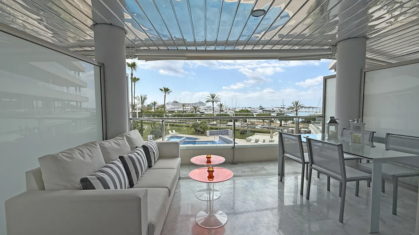 Bright balcony with white sofa, striped pillows, and glass table. Outside, a pool, palm trees, and a harbor with boats are visible.