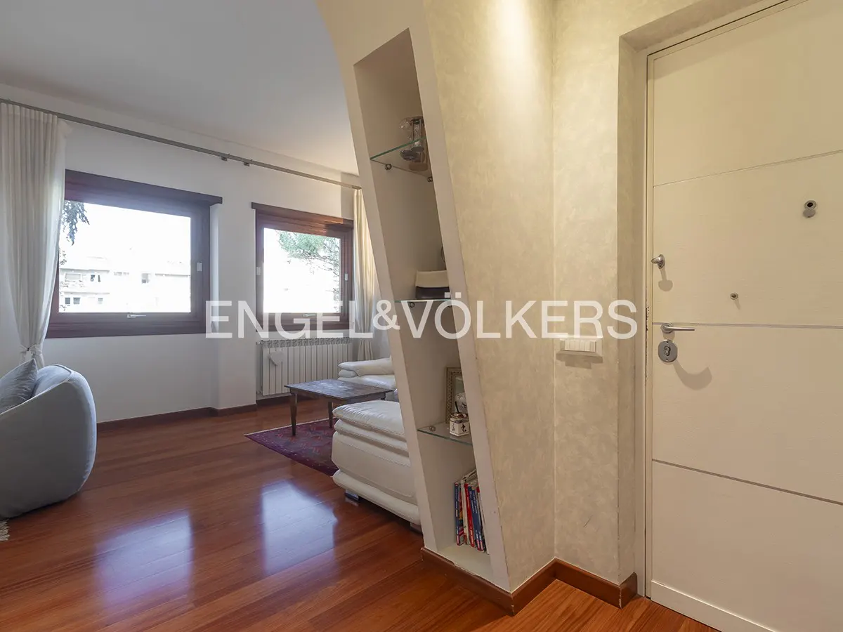 A living room with hardwood floors, windows, a white couch, and a white door. There is a built-in shelf with glass shelves. The Engel & Völkers logo is visible.