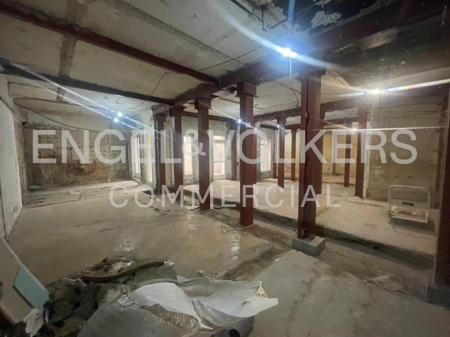 Unfinished commercial space with concrete floors, exposed beams, and red support columns. Engel & Volkers Commercial logo overlay.