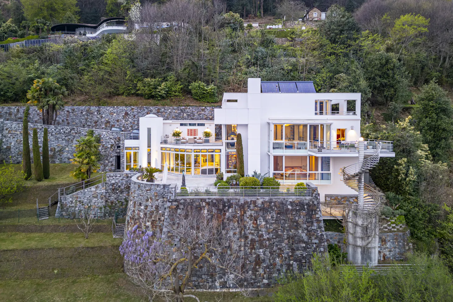 A modern white house with large windows sits on a stone wall, surrounded by green trees and a spiral staircase.