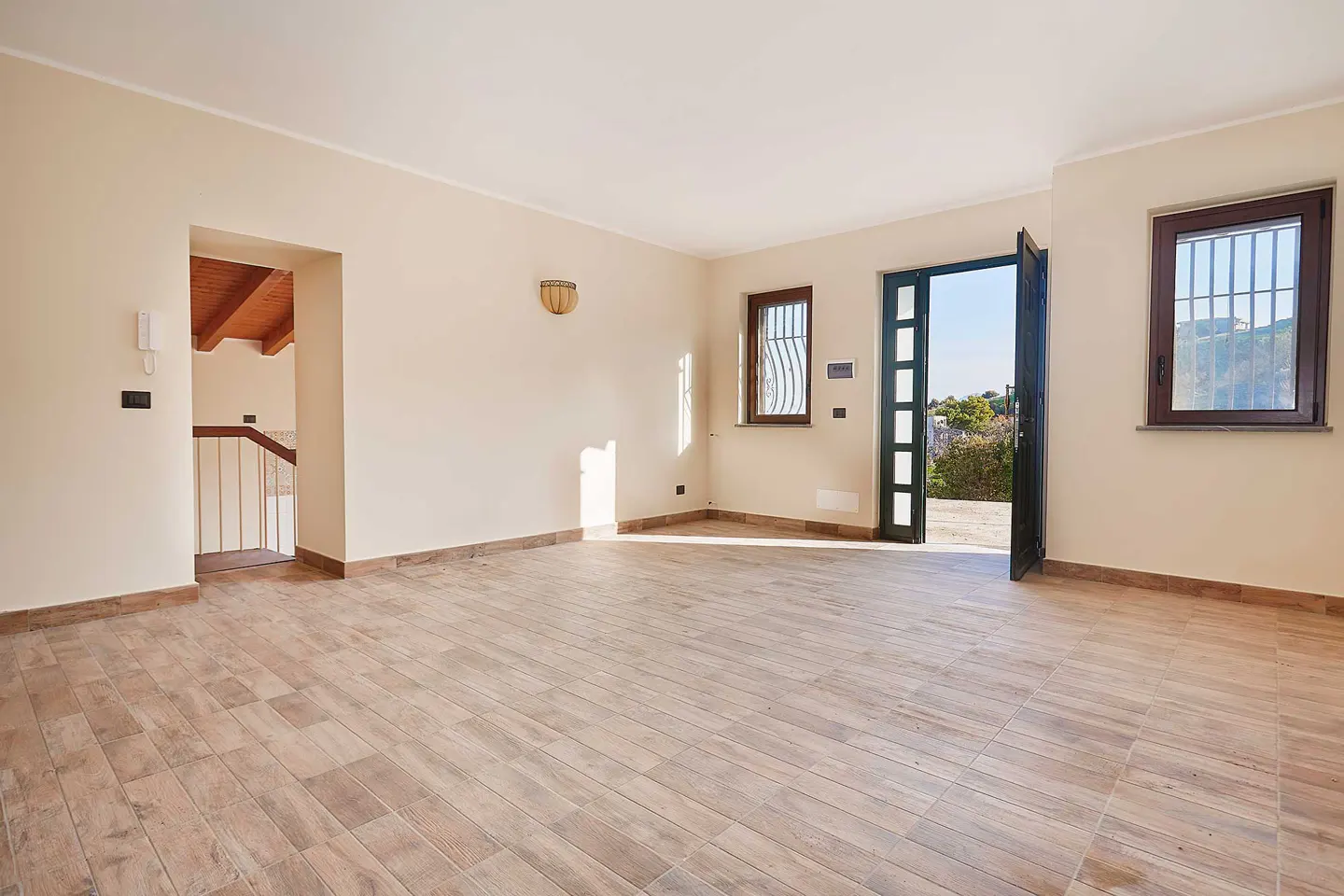 An empty room with beige walls and wood-look tile floors. An open door leads outside, and a doorway shows a staircase.