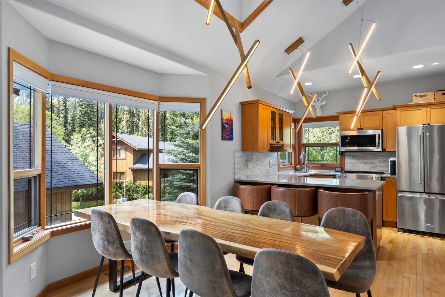 Bright kitchen with wood table, gray chairs, and modern lighting. Large windows overlook trees and a house. Stainless steel fridge and wood cabinets.