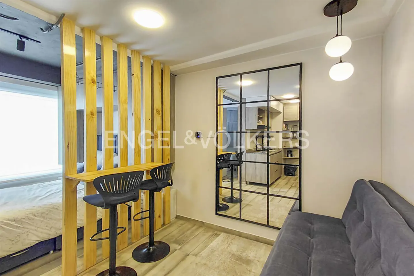 Studio apartment with a wooden slat divider, two black bar stools, and a large grid-patterned mirror reflecting the kitchen. A gray couch is visible.