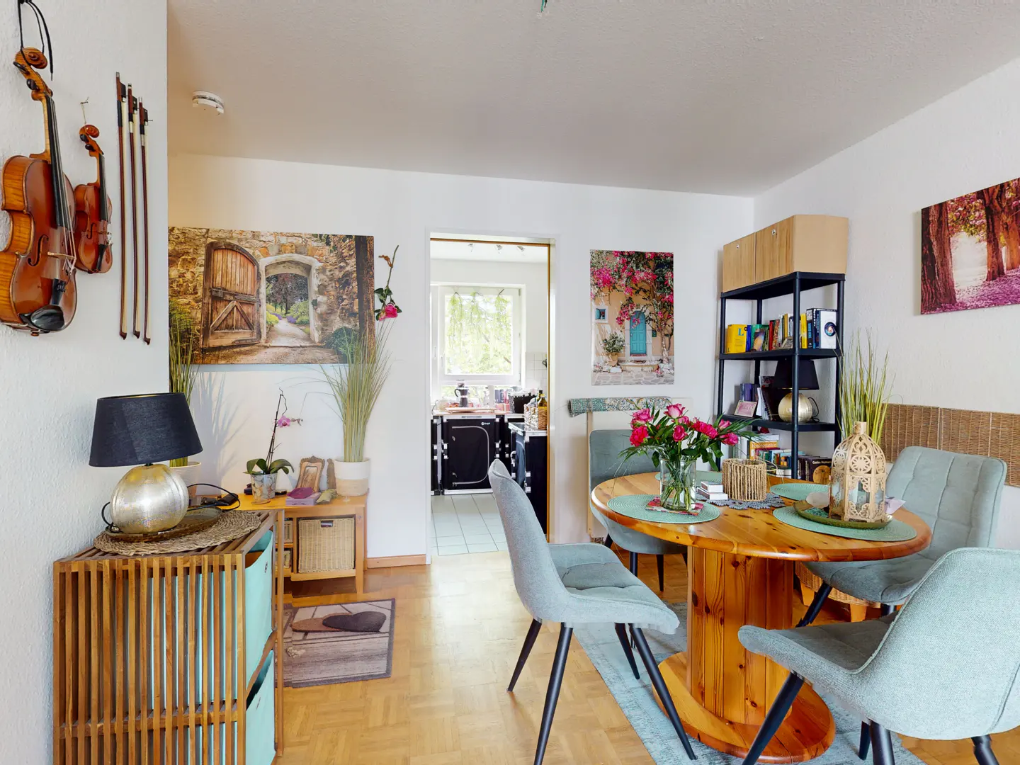 A bright living room with a wooden floor, a round wooden table with four blue chairs, and violins hanging on the wall.