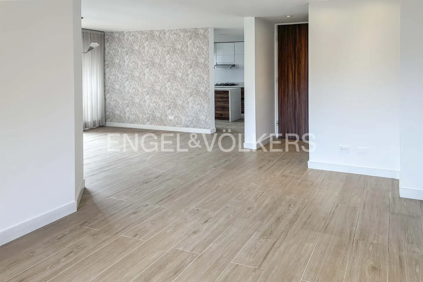 An open-concept living space with light wood floors, white walls, and a kitchen visible in the background. A dark wood door is on the right.