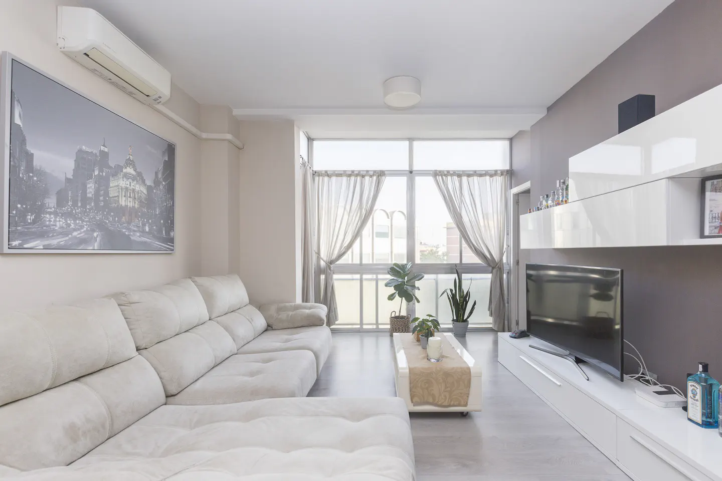 Modern living room with a large beige sectional sofa, white media console, TV, and a city skyline wall art piece.