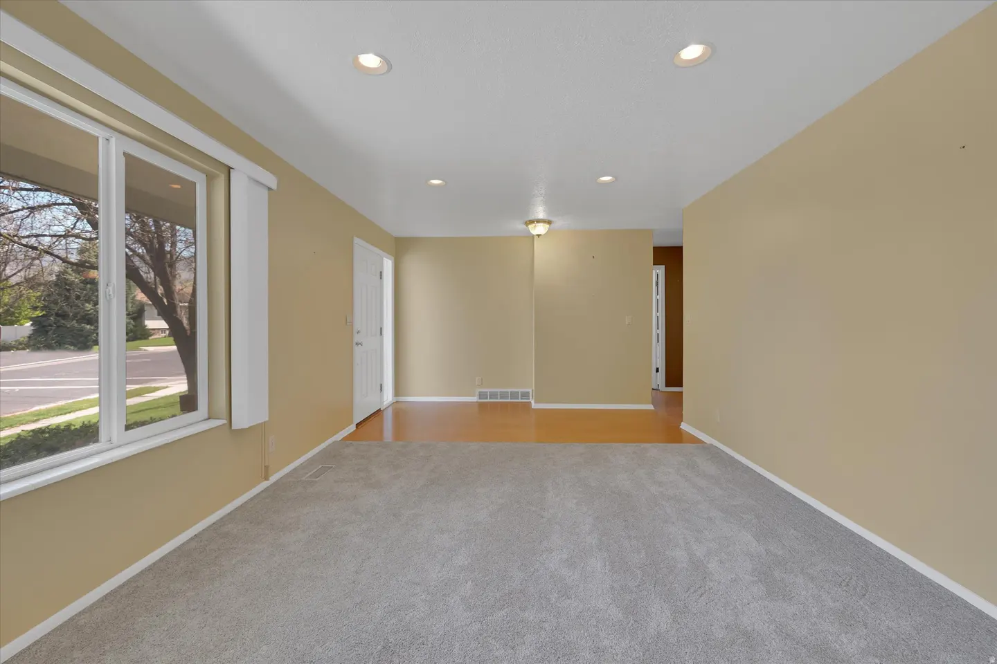 A bright, empty living room with gray carpet, tan walls, and a large window. A white door is visible in the background.